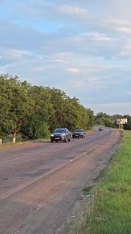 The video shows a rural road with trees on the left and grass on the right. Several cars are driving on the road. The sky is cloudy.