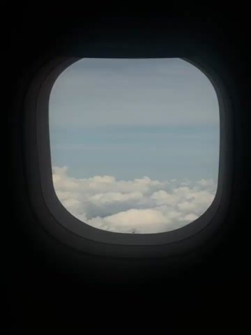 The video shows a view from an airplane window looking out at the sky and clouds. The window frame is dark and circular, with a lighter colored rim. The sky is a pale blue, with fluffy white clouds below. The clouds appear to be at a lower altitude than the airplane. The video is relatively short and appears to be a single, continuous shot. There are no people or other objects visible in the frame besides the window, sky, and clouds.