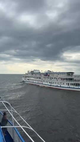 A person stands on the deck of a boat, looking out at a large white ship with multiple decks moving through the water. The ship has red and blue accents and appears to be a river cruise ship, with people visible on its decks. The sky is overcast with dark clouds. The video appears to be shot from another vessel, as the railing of the boat from which the video is taken is visible in the foreground.