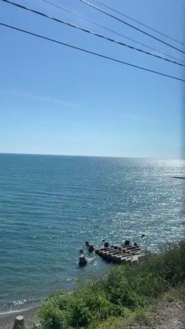 The video pans across a seascape from the perspective of someone looking out a window, possibly on a train or vehicle. The water is a vibrant blue and sparkles under the sunlight. Along the shore, there are sections of concrete barriers and rocky areas. In some shots, there are some green bushes and trees in the foreground. One segment shows a white car parked on the beach near a tent, with two people visible on the concrete structure. Further out, a distant coastline with buildings can be seen. Power lines are visible against the clear blue sky in several frames.