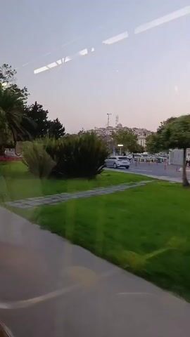The video shows a view from inside a vehicle, looking out a window at a street scene. The window has reflections on it, partially obscuring the view. The street is lined with trees and green grass. Several cars and a motorcycle are visible on the road. There are also pedestrians walking on the sidewalk. A large advertisement billboard is also visible. The sky is overcast, suggesting it might be dusk or early morning. The video is shaky, indicating movement of the vehicle.