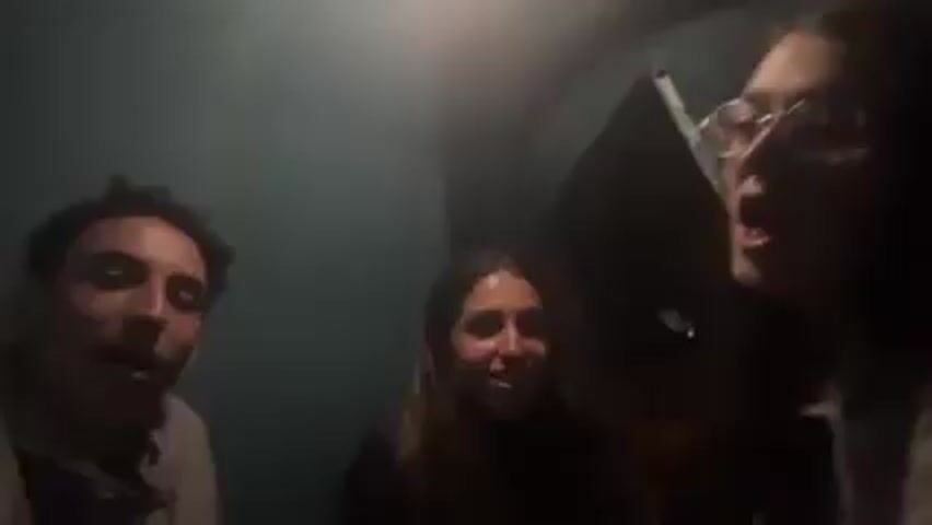 Three young adults are in a dimly lit space, possibly a small room or tent, looking at the camera. On the left, a young man with short, curly hair wears a grey sweatshirt and smiles. In the center, a young woman with long brown hair smiles and laughs. On the right, another young woman with long brown hair and glasses is speaking animatedly. She kisses the woman in the center. A dark object, possibly a piece of equipment or part of the tent, is visible behind them. The background is a solid, dark teal color.