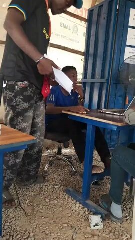 The video shows a small, informal outdoor office setting with people working and waiting. One man in a black shirt and patterned pants stands and appears to be handing a document to a young man sitting at a desk, using a laptop. Another woman with a blue headscarf sits facing the desk, seemingly preoccupied or resting her head on her hand. There's another person, partially visible, using a laptop to the right of the woman.

The office space is characterized by a blue metal structure forming walls, with signs containing phone numbers attached. A small fan is pointed at the workers to keep them cool. The floor is covered with gravel. Other people can be seen in the background, some sitting and some standing, waiting their turn. The lighting seems natural, with an open-air feel.