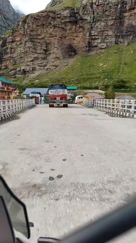 This is a first-person perspective video taken from a vehicle, likely a motorcycle or car, as it travels across a bridge. The bridge has a concrete surface and white metal railings on either side. In the distance, on the bridge, is a large red and white truck with blue accents. Several people are standing near the truck on the left side of the bridge. Behind the truck and people, buildings are visible, suggesting a town or village. To the right of the truck, a white van is parked. The background features a very large, steep, rocky mountain with patches of green grass and vegetation. The sky is overcast. The vehicle appears to be moving forward at a moderate pace.