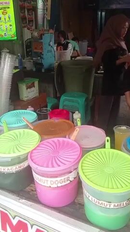 The video shows an outdoor food stall with several large plastic containers of various colored liquids, likely beverages or ice cream ingredients, with their lids on. The lids are brightly colored, with some featuring a ribbed pattern. Labels are visible on some of the containers, with text in Indonesian. In the background, a woman wearing a headscarf is serving a customer. Another person is visible behind a counter with some machinery, possibly for making drinks or ice cream. There are also plastic cups, a fan, and other miscellaneous items on display. The overall setting appears to be a busy street or market.