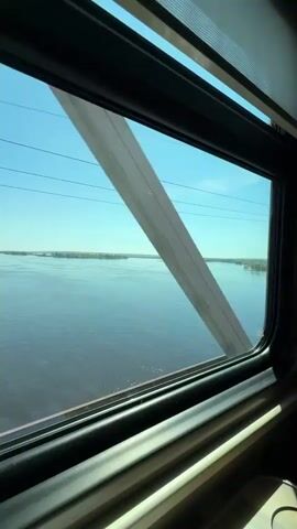 The video shows a bright daytime view from a vehicle, likely a train, looking out a window over a vast body of water. A wide river, possibly the Volga near Vasilyevo, Russia, stretches to the horizon under a clear blue sky, with distant land visible on the far bank. The view is framed by a window, and parts of a metal bridge structure, with visible rivets and beams, are seen through the window. Overhead power lines traverse the sky. Faint numbers "14" and "69" are briefly visible on a vertical beam of the bridge structure. The scene is peaceful and serene, with the water's surface showing gentle ripples.