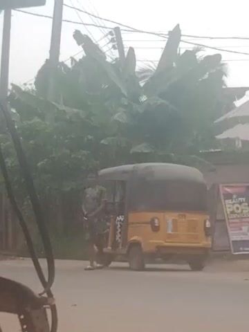 A yellow three-wheeled auto-rickshaw is overturned on a paved road. The vehicle's windshield is shattered. To the left of the auto-rickshaw are several large-leafed plants, possibly banana trees, and power lines. In the background, a poster or sign is visible on a wall. The sky appears overcast or hazy. The orientation of the video is sideways, with the ground appearing on the left side of the frame.