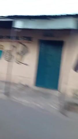 The video shows a blurry scene of a rural area, likely in Africa. The camera is moving, possibly from a car or motorcycle. The landscape is mostly dry and dusty, with sparse vegetation. There are simple, one-story buildings, some made of brick, with corrugated metal roofs. The road appears to be unpaved or poorly paved. There is a motorcycle parked outside one of the buildings. The sky is overcast.