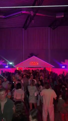 The video displays an indoor scene of a large, dimly lit event space with predominantly purple and pink neon lighting. A high ceiling with exposed metal beams is visible. In the center, a prominent white structure resembling a house or shack with a peaked roof is illuminated by a red neon sign that reads "CAIA." On either side of this structure, palm tree silhouettes with purple backlighting are visible. A dense crowd of young adults occupies the foreground and midground, actively dancing and socializing. Many individuals are holding drinks, and some are holding up their phones. The overall atmosphere is lively and energetic, consistent with a music event or club setting.