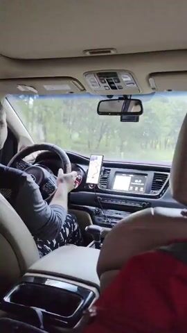 A POV shot from inside a vehicle shows a person's hands on the steering wheel as they drive down a road surrounded by trees. The dashboard features a navigation app displayed on a smartphone mounted on a dash mount, and the car's infotainment system. Traffic is visible ahead, including cars and a large truck. The interior of the vehicle appears to be a minivan, with beige leather seats and a beige headliner. The sky is overcast.