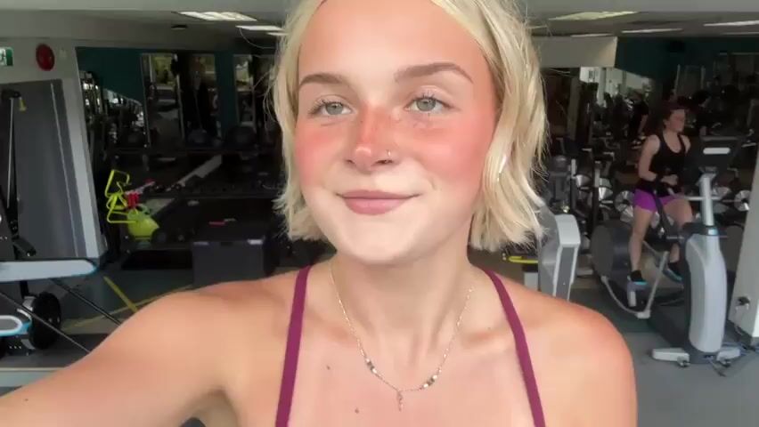 A young woman with blonde hair and a tan is smiling at the camera in a gym. She is wearing a maroon sports bra and maroon leggings. In the background, there are gym machines, including treadmills and stair climbers. The lighting is bright, and the scene is in focus.