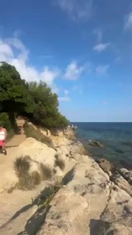 The video shows a rocky coastline meeting the sea under a clear blue sky dotted with fluffy white clouds. The water is a blend of turquoise and darker blues. In the distance, a landmass is visible. A path runs along the coastline, lined with greenery. The scene is natural and serene, presenting a scenic view of the sea and shore.