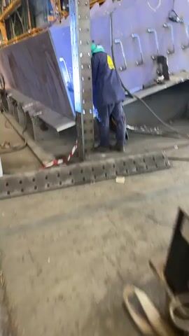 A person in a blue welding jacket and green hard hat is welding a large metal structure. The structure is blue and appears to be a tank or vessel. The person is using a welding torch, which is emitting bright sparks and light. The welding is taking place in an industrial setting, with other machinery and equipment visible in the background. There is a red and white caution tape on the floor in front of the structure. The person's face is not visible as they are wearing a welding mask.