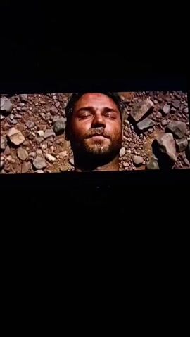 The video shows a person's head lying in the dirt. It then cuts to a shot of a stone building with a door and some vegetation in front of it. Finally, it shows a hand reaching through a field of wheat.