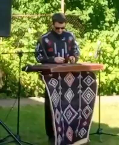 A man is playing a dulcimer outdoors. He is wearing sunglasses and a dark jacket with a patterned design. The dulcimer is on a stand covered with a patterned cloth. There is a speaker to the left of the man, and trees and a fence are visible in the background. The ground is covered in grass.