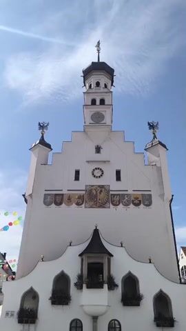 The video shows a wide, low-angle shot of a white building with a tall, multi-tiered tower in the center. The building has a stepped gable facade, with two smaller towers topped with eagle statues on either side of the main tower. Below the main tower, the facade is decorated with several coats of arms and a central mosaic depicting a double-headed eagle. The lower part of the building features a curved facade with arched windows and balconies. Flags or banners are visible in the upper left corner of the frame, appearing colorful and festive. The sky is blue with some wispy clouds, and sunlight is visible, casting some glare on the building. The overall scene appears to be an outdoor shot of a historic building, possibly a town hall or similar public structure, on a bright, sunny day.