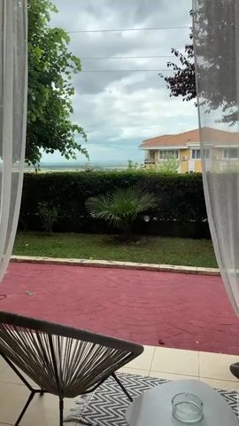 The video shows a view from a patio or balcony through sheer white curtains. In the foreground, a woven chair is partially visible on the left, with a round white table in front of it. The patio floor is covered in red stamped concrete. Beyond the patio, a well-maintained lawn leads to a dense green hedge. In the middle ground, a small palm tree is planted in the lawn. Beyond the hedge, a field or valley stretches into the distance under a cloudy sky. A yellow building with a red-tiled roof is visible on the right side of the frame, behind the hedge. The overall scene suggests a relaxed outdoor setting, possibly a garden or resort area.