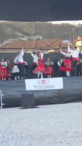 Here's a detailed description of the video:

A group of people are performing a traditional folk dance on an outdoor stage. The dancers are dressed in colorful costumes, with the women wearing long, embroidered red dresses and headscarves, and the men in white shirts, black trousers, and red sashes. They are moving energetically and joyfully, with their arms raised in the air.

The stage is covered in black material and has a banner for "Barros & Barbosa" in front. A speaker sits on the left side of the stage.

Behind the stage, there's a glimpse of a town with buildings with red tile roofs, suggesting a European location. The weather appears bright and sunny. There's a metal fence separating the stage from the audience.

The focus is on a couple in the center of the stage. The woman is twirling, showing off her dress, while the man mirrors her arm movements. An accordion player is visible in the background, likely providing the music for the dance.
