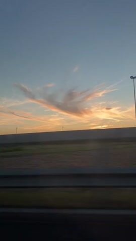 The video shows a view from inside a vehicle looking out at a sunset. The sky is a clear blue with wispy, pink and orange clouds. In the distance, there is a flat landscape with fields and a long, dark barrier. The sun is setting on the horizon, casting a warm glow. The video is shot from a moving vehicle, indicated by the slight blur and the reflection of the vehicle's interior on the window. There are no people or discernible objects in the foreground.