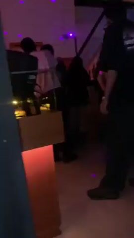 The video shows a dimly lit indoor scene, possibly a bar or club, with several people present. There's a man in the foreground wearing a black t-shirt, dark pants, and shoes. Several other people are visible in the background, some standing near a lit countertop or bar area. One person is wearing a white shirt. Blue lighting and disco lights are apparent. The video's perspective seems to be from behind or to the side of a doorway. The background contains details like hanging decor and what appears to be a partial view of another level.
