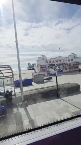 The media file shows a view of the Grand Pier in Weston-super-Mare, United Kingdom, from what appears to be inside a vehicle. The scene is set on a sunny day with scattered clouds. The Grand Pier, a prominent white building with red lettering and domes, stands in the background, extending out towards the sea. People are visible walking on the pier and on the paved promenade in front of it.

In the foreground, the paved promenade is laid out with light-colored square tiles. A grey stone wall runs along the edge of the promenade, separating it from a lower paved area. There are several objects visible, including a blue container, trash bins, and a lamppost with a speed limit sign indicating "30". The sea and the sandy beach are visible behind the pier. The overall atmosphere appears to be a bright and pleasant day, with people enjoying the seaside location. The view suggests the media was captured during daylight hours.