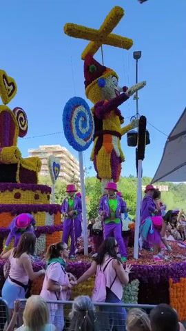 A flower parade float depicts a large, brightly colored clown figure and a cake, adorned with various flowers. Several performers in purple and green outfits with pink hats dance on the float. Spectators, including women and children, are visible in the foreground, some holding up cell phones to record the event. The sky is clear and blue.