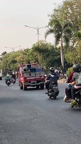 The video shows a busy street scene. In the foreground, several motorbikes are traveling on a paved road. On the right side of the road, there are palm trees and other greenery. In the background, there is a red truck with items loaded onto its bed, and several other vehicles, including motorbikes and what appears to be a larger truck. The sky is overcast, suggesting it may be daytime. The overall impression is one of moderate traffic on an urban or suburban road.