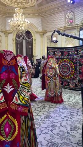 A group of women in colorful traditional clothing are dancing in a room with ornate decor. One woman in the foreground, wearing a red top and a brightly patterned skirt, performs a graceful dance move with her arms. In the background, other women in similar attire are also dancing, some with their faces covered. A tapestry with a circular design hangs on the wall. The room is lit by a large chandelier and features patterned carpets and tall windows. There are also people in the background who appear to be observing the performance.