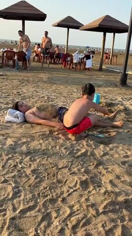 A young boy, wearing red swim trunks, is playing in the sand on a beach. He is crouching and holding an orange plastic shovel. In front of him, another boy is lying on his back in the sand, partially covered by sand up to his torso. The boy on the ground has a white pillow under his head. In the background, there are beach umbrellas, chairs, and tables, with other people in the distance. The sun is shining, indicating it is daytime.