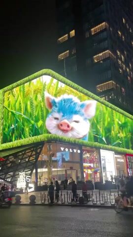 The video shows a large LED billboard at night in a city. The billboard displays a cartoon pig with blue hair and a smiling face surrounded by a field of green wheat. Below the billboard are storefronts, including Volvo and Dreame, with people walking around. A car passes in front of the camera. A tall building stands in the background.