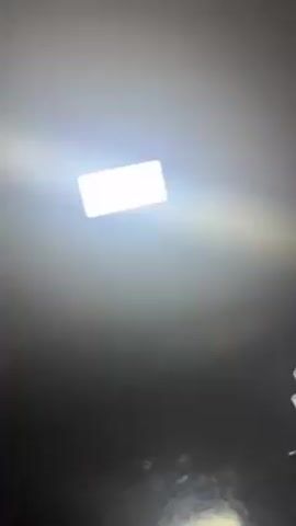 The video depicts a square light fixture against a dimly lit ceiling, with a wooden beam briefly entering the frame from the top left. There are unclear objects in the lower part of the frame. The camera appears to be held at a low angle, pointed upwards at the light.