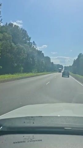The video shows a view from inside a vehicle driving on a two-lane highway. The road is paved and flanked by trees on both sides. The sky is blue with some white clouds. Several vehicles are visible on the road ahead, including a truck and a car. The truck passes the camera vehicle at 00:02. The camera is pointed forward, and the dashboard of the vehicle is partially visible at the bottom of the frame. The overall impression is of a sunny day driving on a rural highway.