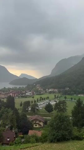 The video pans across a cloudy, mountainous landscape with a body of water and a small town. The foreground shows several houses with red-tiled roofs and some greenery. The middle ground features a town with multi-colored buildings, a park-like area with trees, and a parking lot filled with cars. Beyond the town, a large lake or bay is visible, surrounded by steep, forested mountains. The sky is overcast with dramatic, gray clouds, and there is a hint of yellow light breaking through in the distance. The overall impression is of a somewhat moody, atmospheric day in a scenic, European-style village.