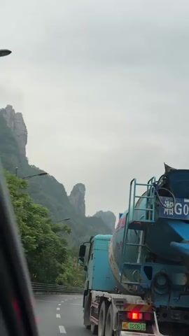 The video shows a blue cement mixer truck driving on a wet highway next to a guardrail. There are cars driving in the opposite direction. In the background are tall, rocky mountains shrouded in mist and fog, with green trees and vegetation. The sky is overcast. The camera is moving at the same pace as the truck, looking out of the window of a vehicle. The truck has Chinese characters and numbers on it.