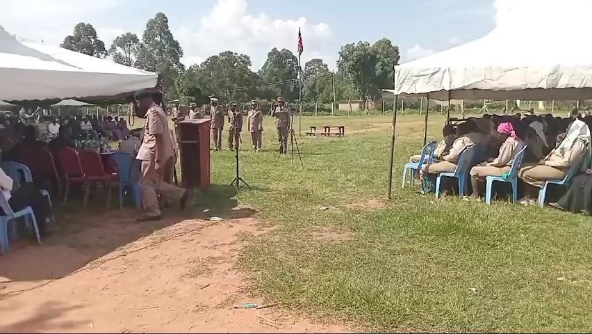 The video captures an outdoor ceremony or gathering in Lwandeti, Kenya, on a bright, sunny day. The scene is set on a grassy field with trees and a building in the background. Two large white tents with green and red accents provide shade for an audience, predominantly women, seated on plastic chairs.

In the foreground, several men and one woman, all dressed in tan uniforms with matching hats, stand in a line facing the audience. These individuals appear to be government officials or community leaders. A wooden podium with a microphone is positioned in front of them. Throughout the video, various uniformed individuals approach the podium, salute, introduce themselves by name and title (e.g., "Assistant Chief Kiliboti Sub-location," "Chief Matulu Location"), and then move to sit at a table under a tent or back into the line.

A man in a green shirt and jeans is seen operating a camera or phone, filming the event near the podium. Another man is seen setting up or adjusting a microphone on a stand. The atmosphere appears formal and organized, with the officials performing a protocol or presentation. The audience remains seated and observant. In the background, a Kenyan flag is visible on a flagpole.