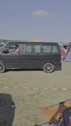 The video begins with a dark van with tinted windows driving slowly on a grassy field. The driver is visible and wearing a hat. The van has custom rims. The scene then shifts to a wider shot showing a field filled with cars and tents, suggesting a festival or camping event. Several cars are parked haphazardly, and various tents and camping shelters are set up. In the background, there are trees and a blue sky with clouds. Another shot shows a yellow van, a dark sedan, and more tents. A black car's rear is prominently featured in the foreground, with its roof antenna visible. The video concludes with a view of a tent setup with chairs and tables, and two people sitting under a canopy. One person is wearing a pink shirt and shorts, while the other is facing away from the camera. The overall environment is outdoors, with a bright, sunny sky.