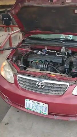 Here is a description of the video:

The video shows a red Toyota Corolla with its hood open at a gas station. A black hose is connected to the car's engine compartment from a white gas pump. The car has a Kogi license plate reading LKJ-313T. A man in a black shirt and blue jeans walks away from the car and toward the pump. Behind the car, a yellow truck is parked. Another man is visible sitting on a red barrier near the truck.