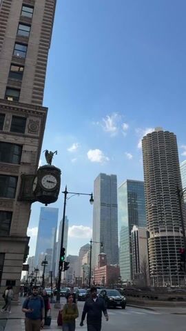 The video shows a streetscape in a large city, with multiple high-rise buildings visible against a clear blue sky. In the foreground, pedestrians walk across a crosswalk at a street intersection. There are cars on the road, stopped at a red light. Several notable buildings are visible, including Marina City towers, a dark-colored skyscraper, and the Trump International Hotel and Tower. A street sign indicates Wacker Drive. The video pans upwards, focusing on the tall buildings.