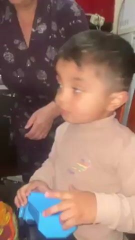 A toddler is sitting in a wooden chair, holding a blue toy in his hands. He is wearing a long-sleeved tan shirt with a colorful design. To the left of the toddler, an older woman is standing, wearing a dark-colored floral patterned robe. The toddler opens his mouth wide, looking directly at the camera. A red cabinet is visible behind the woman, and a microwave is to the left. The video appears to be filmed indoors in a kitchen setting.