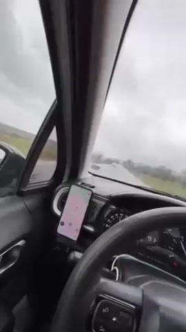 The video shows a car's interior, specifically the driver's side, while driving on a road. It's a gray, overcast day, and the windshield wipers are active. A phone mounted on the dashboard displays a navigation app with a visible route. The steering wheel is black with several buttons. The car's speedometer shows a speed around 93-99 km/h. The outside environment includes fields and trees. A vehicle can be seen ahead on the road. Music is playing in the background.