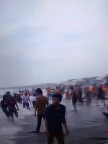 The video shows a crowded beach with many people walking in shallow water and on the wet sand. Several colorful umbrellas are set up on the beach. There are also some motorcycles and various objects scattered around, possibly merchandise or personal belongings. The sky is overcast. People appear to be enjoying themselves, with some children playing in the water. The scene is chaotic and busy, with a lot of movement and activity.