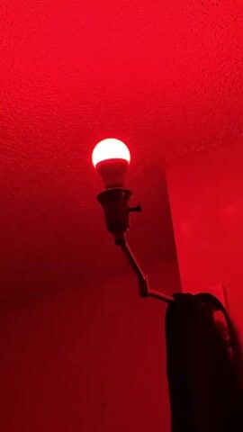 A dim, red light illuminates the scene. A lamp with a visible white lightbulb is angled upwards towards the ceiling. The ceiling and the surrounding wall are also bathed in red light. The lamp’s cord and a dark object are partially visible.