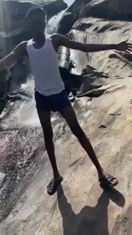 The video shows a dark-skinned person standing in front of a small waterfall. The person is wearing a white tank top and blue shorts, and sandals. They are standing on a large rock, with the waterfall in the background. The person is extending their arms, and smiling. The sun is shining, and the sky is blue. The environment is rocky and natural.
