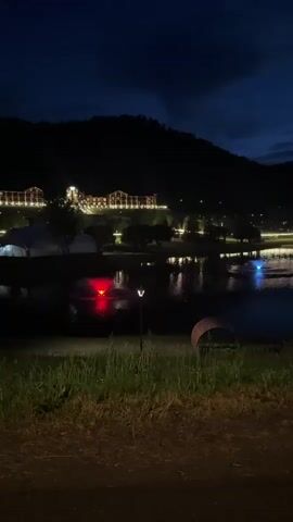 This is a vertical, nighttime video of a scene by a body of water. In the foreground, there is a patch of grass with some shadows and a dark path. A curved, metallic object, possibly a bench or sculpture, is visible on the right. Further back, there is a red illuminated object, possibly a car's taillights, and a small lamp post. The body of water reflects lights from buildings and trees on the opposite shore and across the water. On the far shore, there are buildings illuminated with many lights, situated on a hillside. The sky is dark blue with some clouds. The overall impression is a serene, nighttime landscape with man-made structures and natural elements.