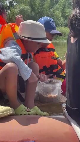 The video shows two young boys on a boat, likely on a river, surrounded by lush green trees. Both boys are wearing bright orange life vests. The boy on the left is wearing a white hat and a light blue shirt, and he is looking down at a white bucket filled with what appears to be fish bait. He is holding a small tool, possibly a fishing hook, in his right hand. The boy on the right is wearing a blue baseball cap and a yellow shirt. He is looking at the bucket and holding a spoon or similar utensil, seemingly also engaged with the bait. The water is calm and a light brown color. The scene suggests a fishing activity.