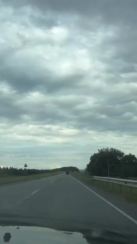 The video captures a forward-facing view from inside a vehicle traveling on a paved road under a cloudy sky. The road appears to be a highway with multiple lanes, marked by white lines. To the left of the road, there is a grassy shoulder and a line of trees. To the right, there is a dense forest and a metal guardrail. The sky is filled with heavy, grey clouds. A few cars are visible in the distance, also traveling in the same direction. The overall scene suggests a journey through a rural or semi-rural area.