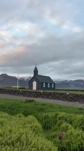 This image captures the iconic black church of Búðir, Iceland, set against a dramatic natural backdrop. The church itself is a small, dark-colored wooden structure with a steep, dark roof and a slender steeple topped with a cross. It features several small, white-framed windows and a prominent white front door, which provides a stark contrast to its dark siding.

In the foreground, lush green grass and various low-lying plants, possibly ferns or similar foliage, stretch across the bottom of the frame. A dark, rugged stone wall runs horizontally through the middle ground, separating the grassy area from a dirt or gravel path that leads towards the church. To the left of the church, a tall, white flagpole stands upright.

The background is dominated by a majestic range of mountains, some of which still bear patches of snow on their peaks, indicating a cool climate. The sky is largely overcast with grey clouds, creating a soft, diffused light over the entire scene. The overall impression is one of peaceful solitude and remote beauty. No people or visible text are present in the image. The time of day appears to be daytime, possibly under an overcast sky.