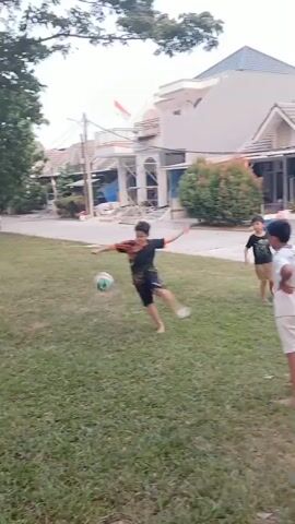 Two young boys are playing soccer in a grassy park. The boy on the left is wearing a black t-shirt and shorts, and the boy on the right is wearing a white t-shirt with the number 9 on the back and white shorts. The boy on the left is kicking the ball towards the boy on the right, who is in a ready stance to receive it. The background shows a residential area with houses and trees. The sun is setting, casting a warm light on the scene.
