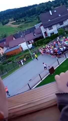A large group of runners are gathered on a paved road, facing forward, ready to begin a race. The runners are of various ages and genders, and they are wearing colorful athletic clothing. Some runners are standing in the front, while others are behind them. The road is lined with houses and trees, and there is a grassy area on the right side of the road. In the foreground, a child's hand with a temporary tattoo is visible, resting on a wooden surface. The sky is overcast.
