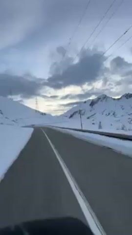 The video shows a POV shot of a road winding through a snowy mountain landscape. The sky is cloudy, with hints of sunlight breaking through. There are power lines visible above the road. The road itself is asphalt, with a white line dividing the lanes. As the camera moves, it passes a small creek and a wooden guardrail on the side of the road. The mountains are covered in snow, with a few trees scattered across the landscape.